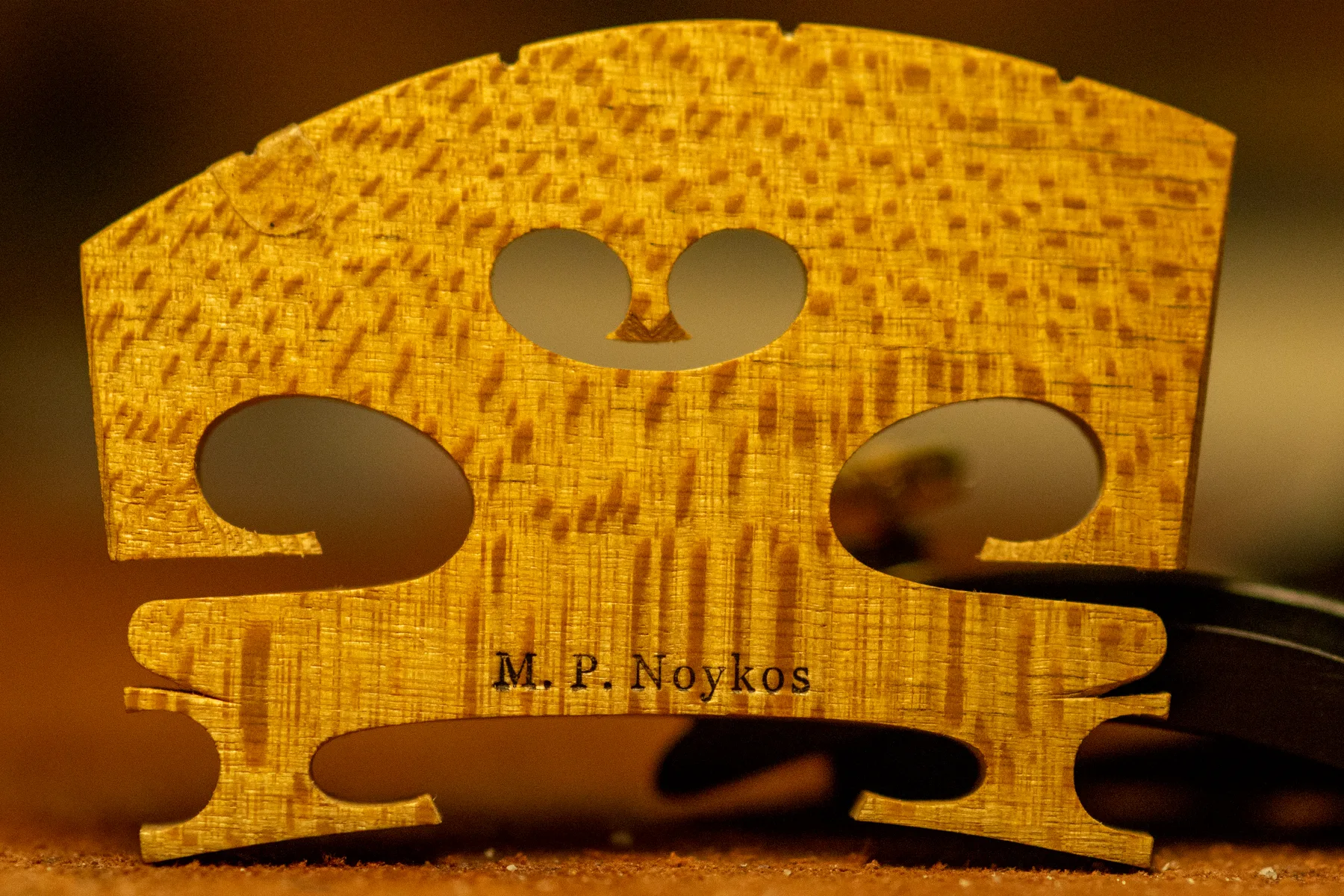 A hand-carved, backlit maple violin bridge stamped “M. P. Noykos,” glowing amber.