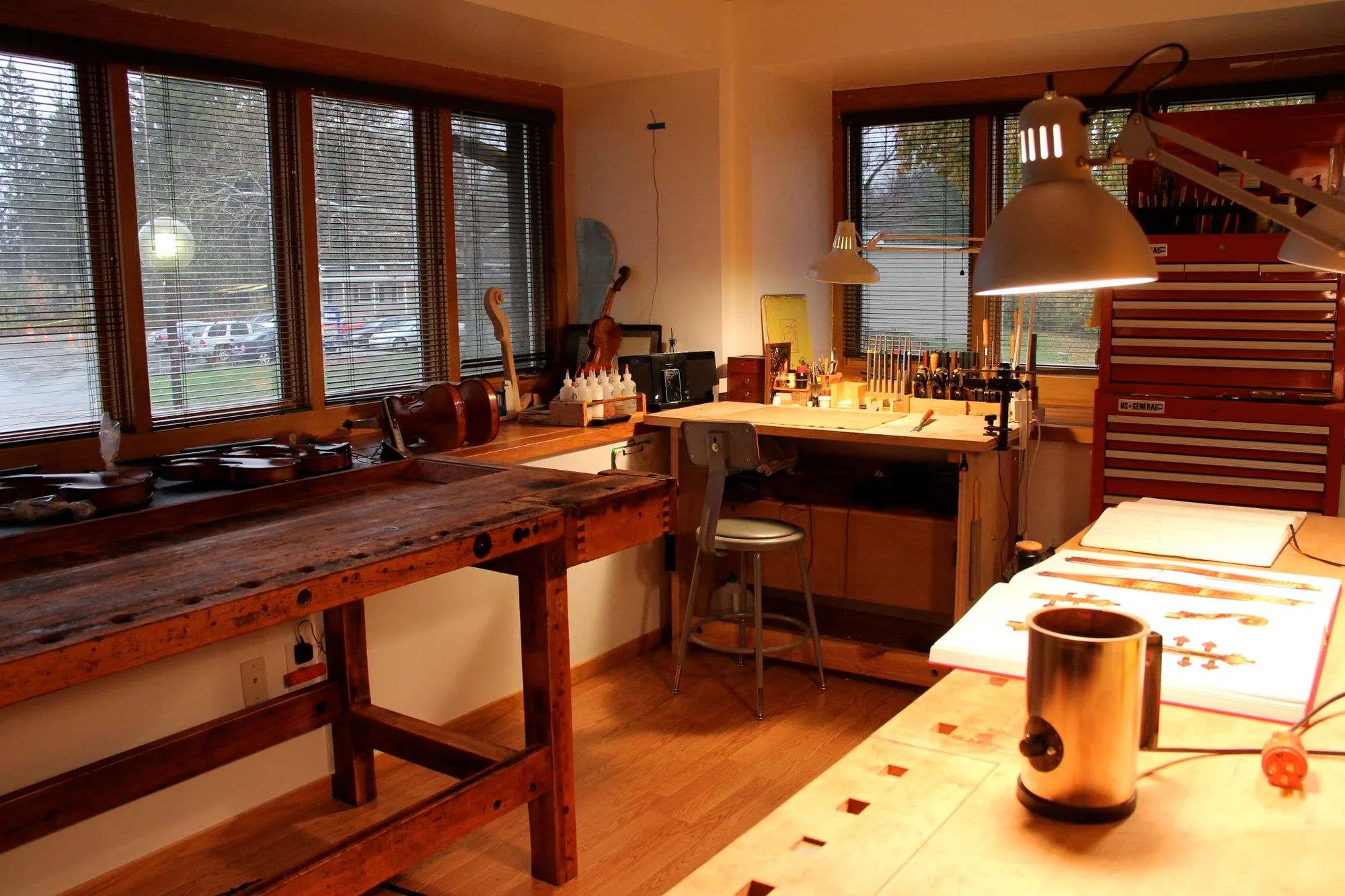 The Grand Rapids Violins workbench at last light — two reclaimed-wood benches, instruments hung in the window, a shop lamp, late-afternoon window light.